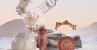 Trije Premiers Rayons izdelki praznične kolekcije L'Occitane obdani s sceno snega, kamna in belih rož ob sončnem vzhodu