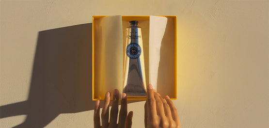 Krema za roke Karite za suho kožo je položena v odprto darilno škatlo L’Occitane, ki jo držijo ženske roke