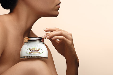 Ženska čepi in na kolenu drži Mandljevo koncentrirano mleko L'Occitane