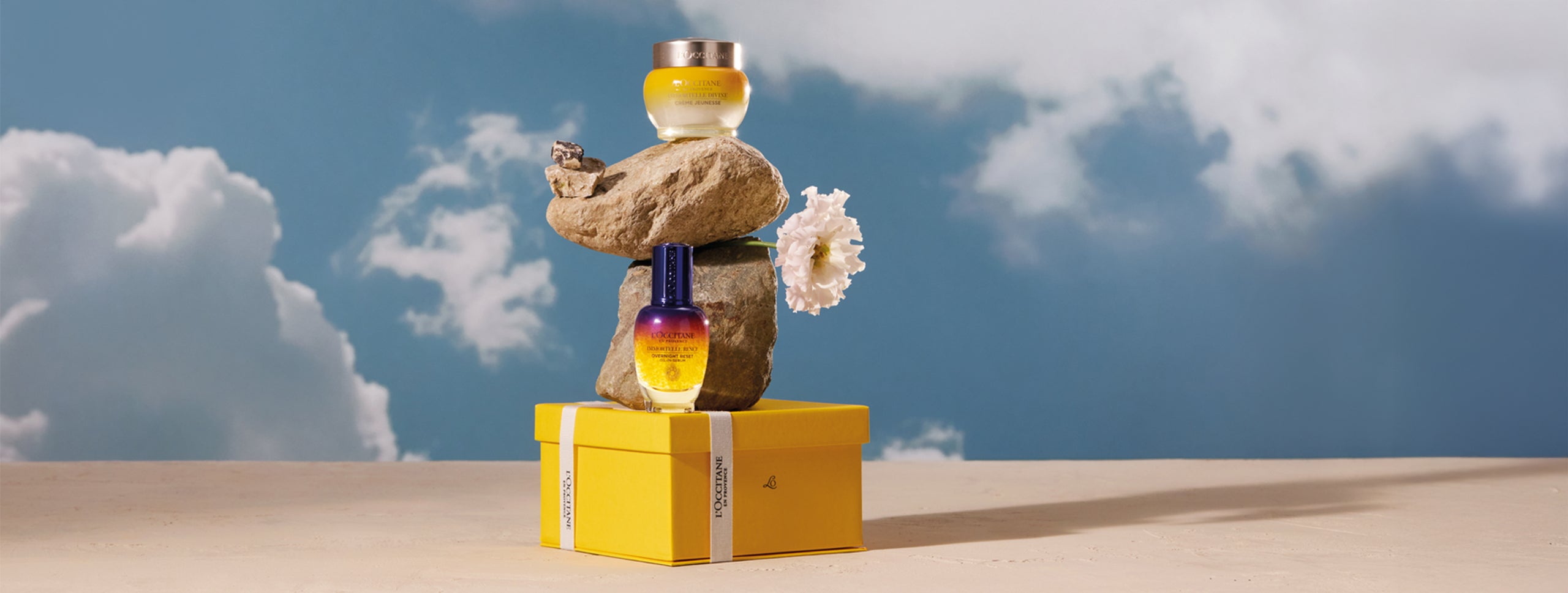 L’Occitane Booster za nočno nego obraza Immortelle Reset n a rumeni darilni škatli, na njej kamna in roža, na kamnu L’Occitane Božanska krema za obraz, v ozadju nebo