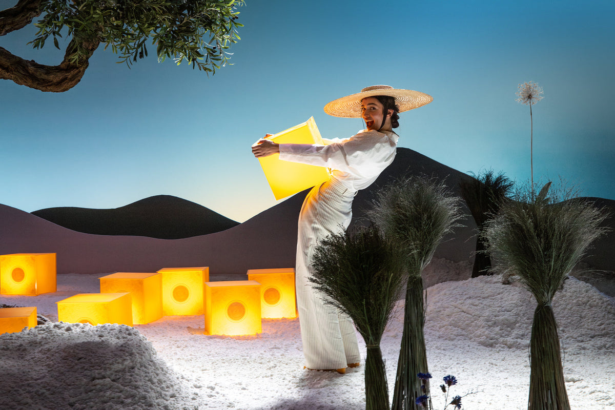 Ženska s klobukom, ki stoji in v rokah drži rumeno darilno škatlo L'Occitane.