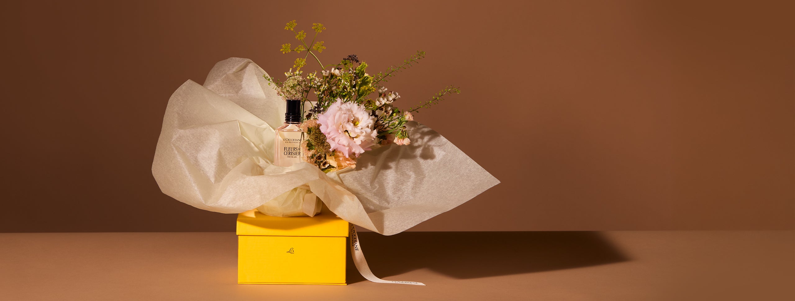 Na rumeni darilni škatli L’Occitane sta postavljena šopek in steklenična dišave L’Occitane   