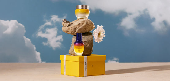 Na rumeni darilni škatli L'Occitane sta postavljeni steklenički Boosterja za nočno nego obraza Immortelle Reset in Božanske kreme – L'Occitane – v ozadju je modro nebo 