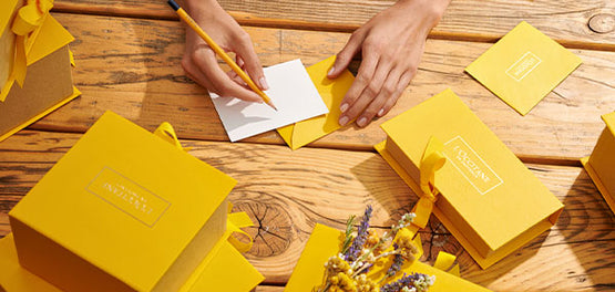 Pisanje voščilnice za rojstni dan na mizi, polni darilnih škatel L’OCCITANE v rumeni barvi 

