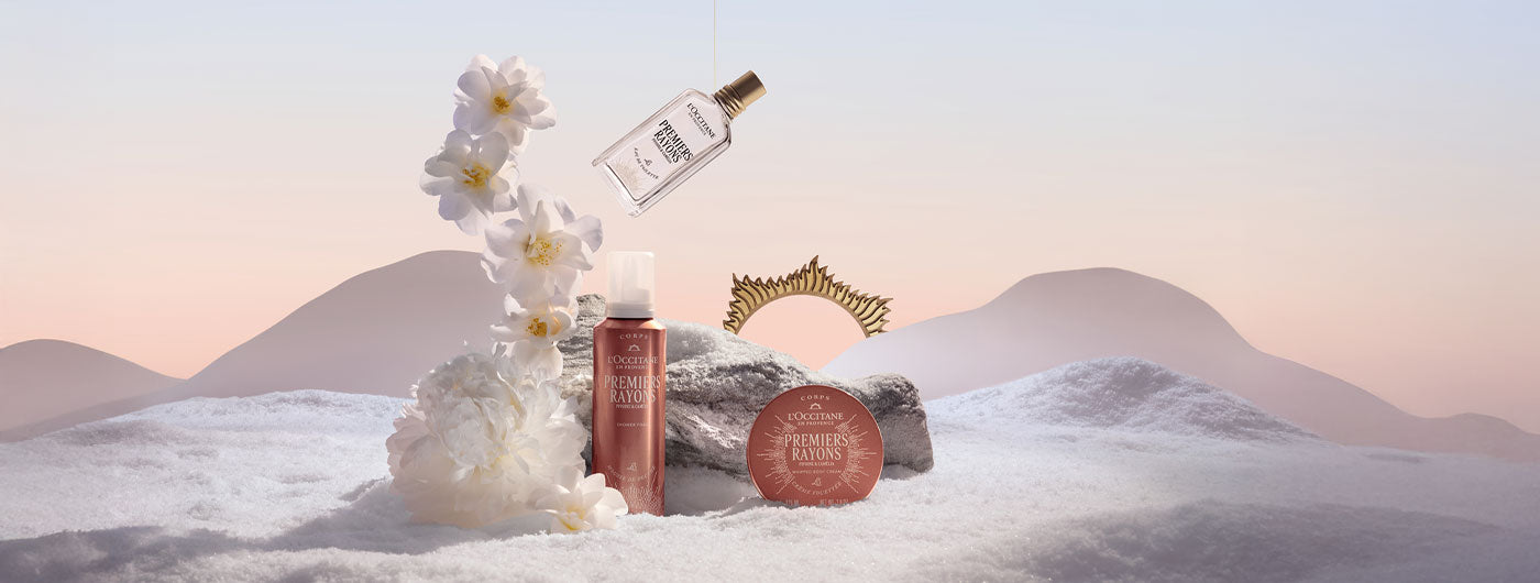 Trije Premiers Rayons izdelki praznične kolekcije L'Occitane obdani s sceno snega, kamna in belih rož ob sončnem vzhodu