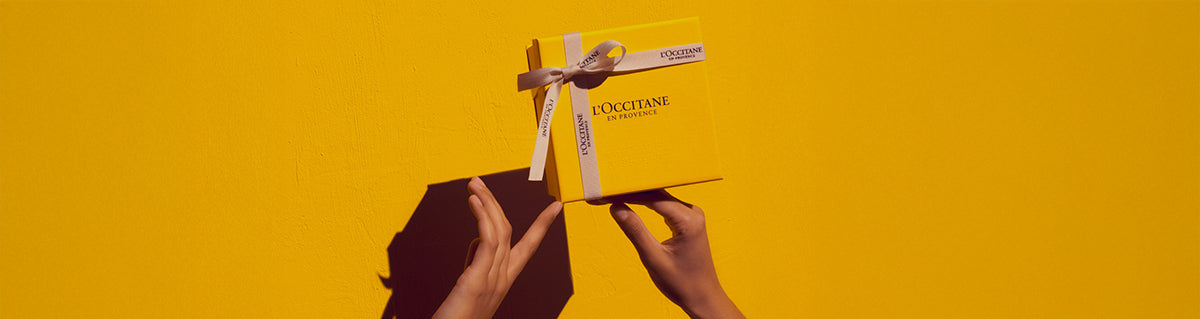 Roke držijo darilno škatlo l’Occitane – rumena škatla na rumenem ozadju 