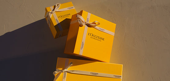 Tri darilne škatle L’Occitane z belim trakom, postavljene na bež podlago.