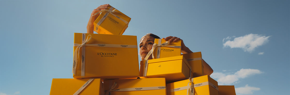 Ženska gleda izza L’Occitanovihe darilnih škatel, naloženih enoa na drugo, za njo soje modro nebo in oblaki 