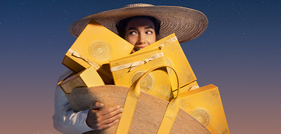 Ženska s klobukom drži v rokah L’Occitanove darilne škatle v rumeni barvi.