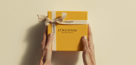 Ženske roke držijo L’Occitanovoe darilno škatlo v rumeni barvi z belim darilnim trakom, zavitim okrog nje. 