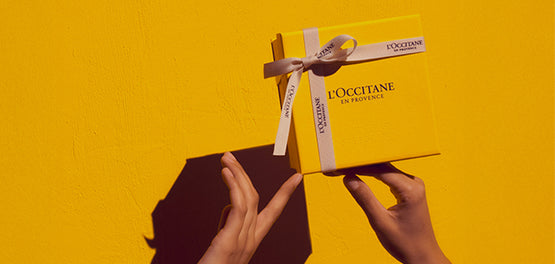 Ženske roke s konicami prstov držijo L’Occitane darilno škatlo v rumeni barvi, na rumenem ozadju 