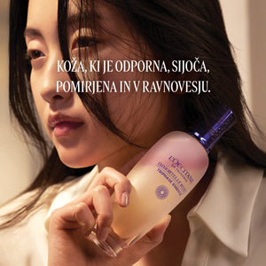 Ženska, ki ob obrazu drži izdelek L’Occitane Trifazna esenca za obraz - Immortale Reset kolekcija