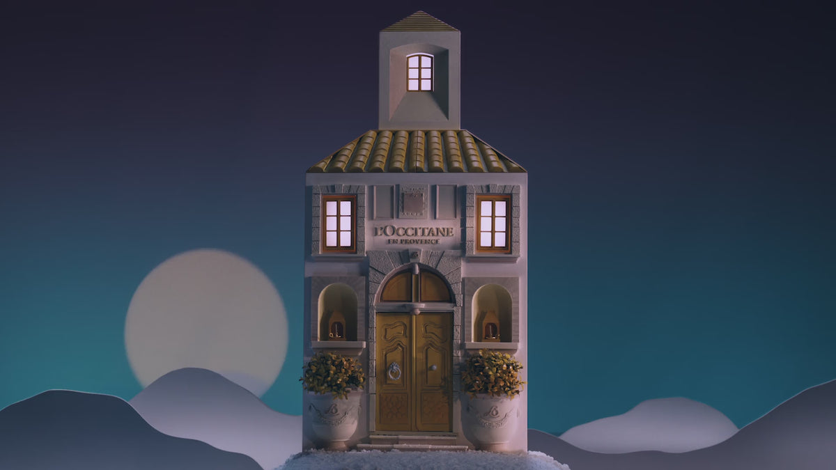L'Occitane praznični adventni koledar v obliki hiše. Vanj pogleda ženska v beli obleki in čez okno vidi različne praznične izdelke.